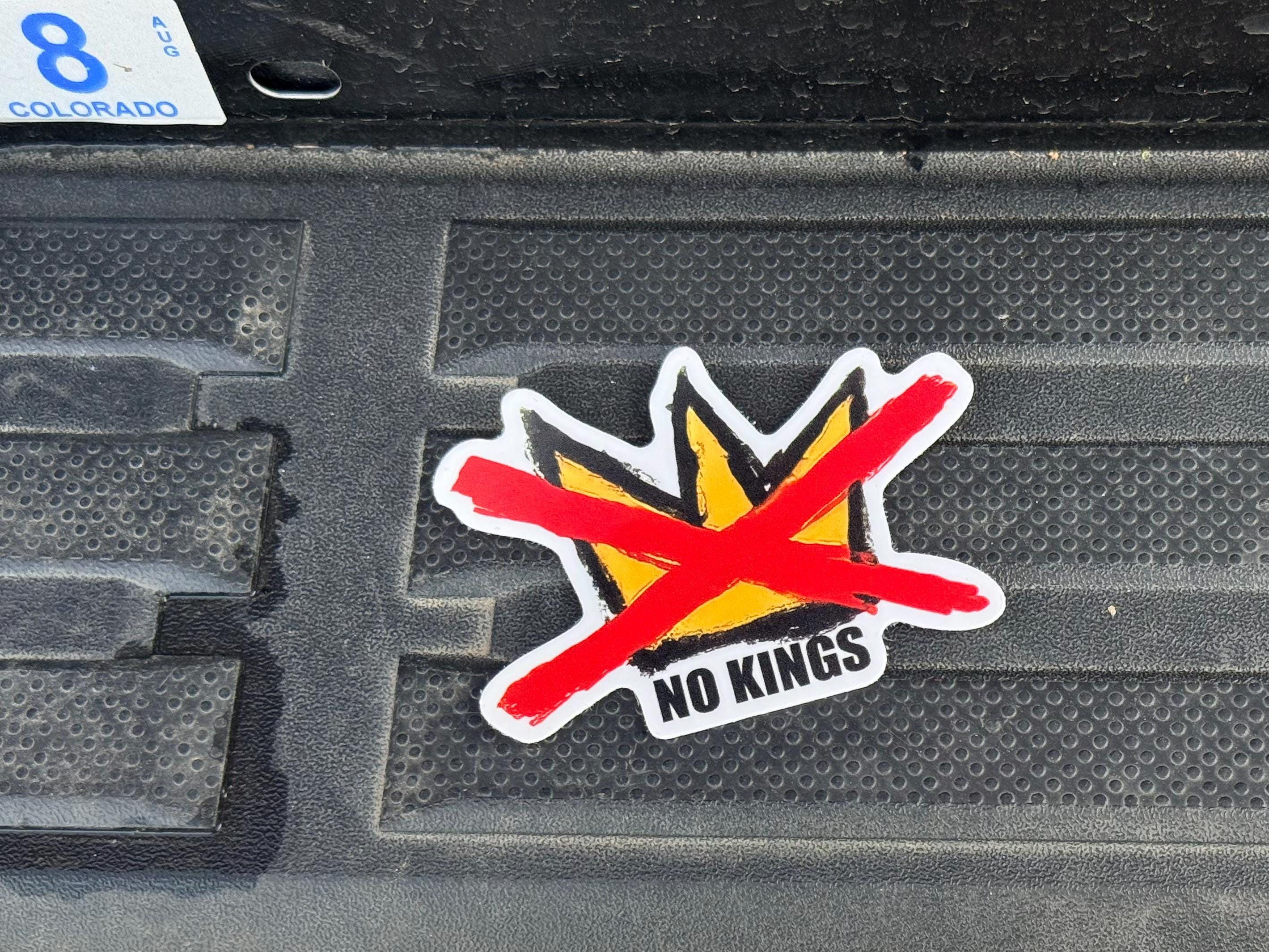May include: A sticker with a yellow crown design crossed out by a red line. The sticker has the words "NO KINGS" in a white font. The sticker is on a textured black surface.