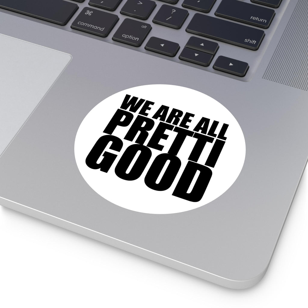 May include: A white, circular sticker with the phrase "WE ARE ALL PRETTI GOOD" in bold, black, sans-serif font. The sticker is placed on a silver laptop, with the keyboard visible in the background.