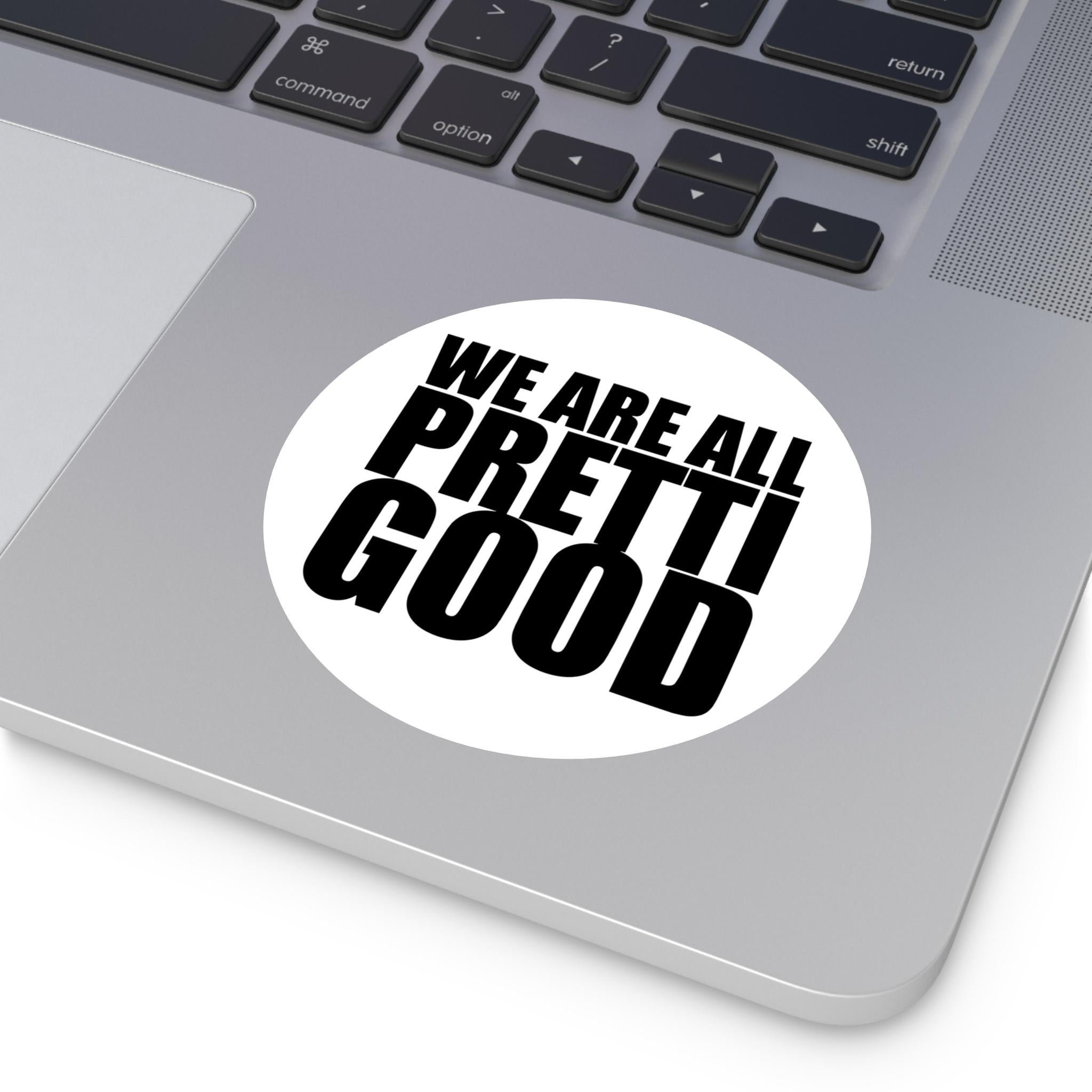 May include: A white, circular sticker with the phrase "WE ARE ALL PRETTI GOOD" in bold, black, sans-serif font. The sticker is placed on a silver laptop, with the keyboard visible in the background.