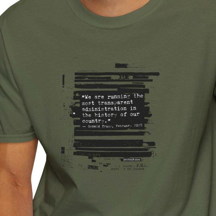 May include: Olive green t-shirt with a black text design. The text is a quote, partially obscured by horizontal black bars. The person in the image is wearing blue jeans. The shirt is a short-sleeved, crew neck style.