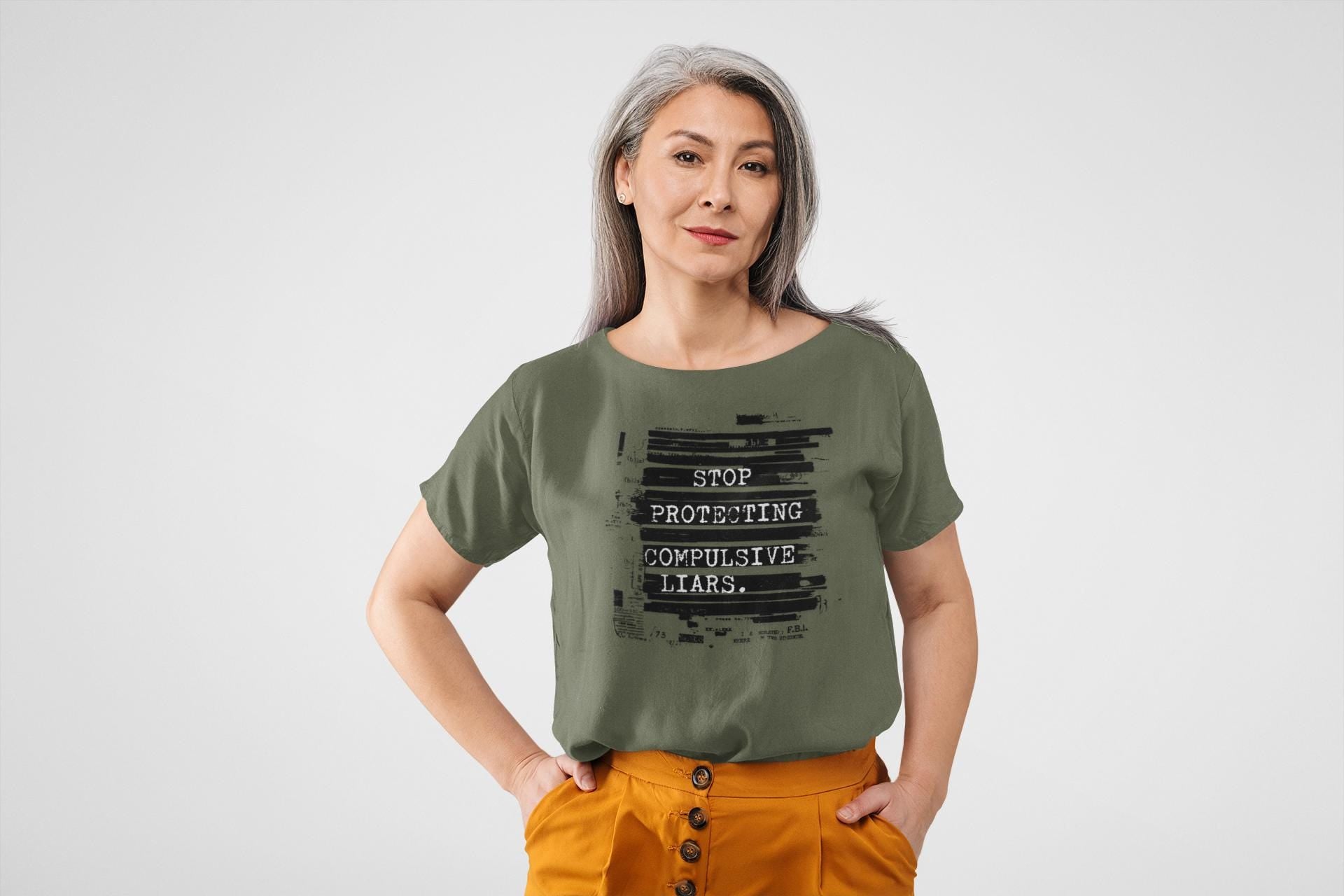 May include: Olive green t-shirt with the text "STOP PROTECTING COMPULSIVE LIARS" in a black, typewriter-style font. The model is wearing orange pants. The shirt has a round neck and short sleeves.
