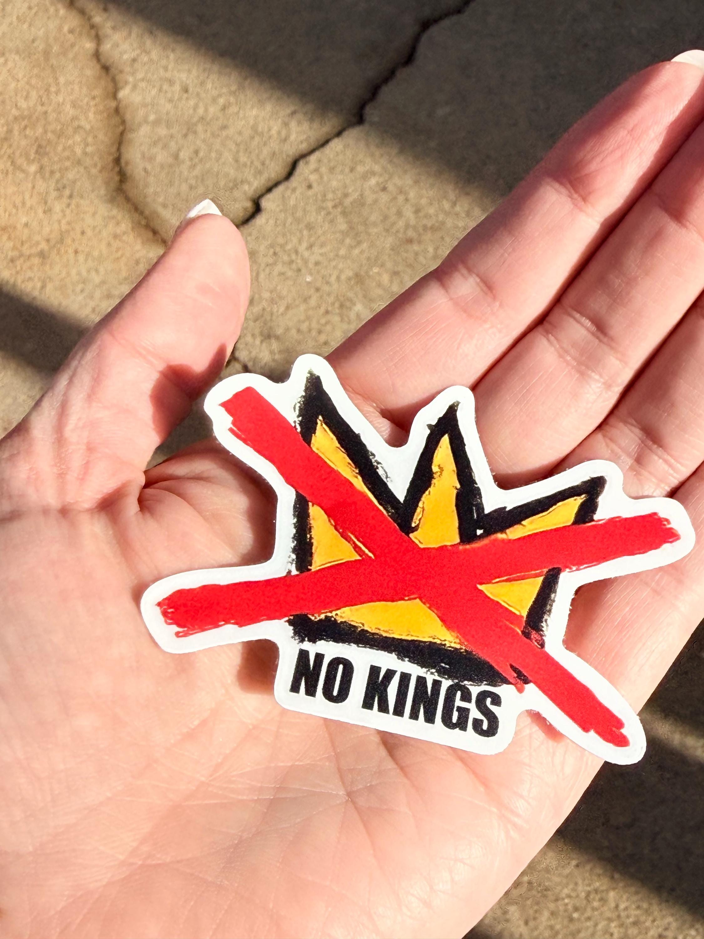 May include: A sticker featuring a yellow crown design with a red slash through it. The words "NO KINGS" are printed in black below the crown. The sticker has a white border and is held in a hand.