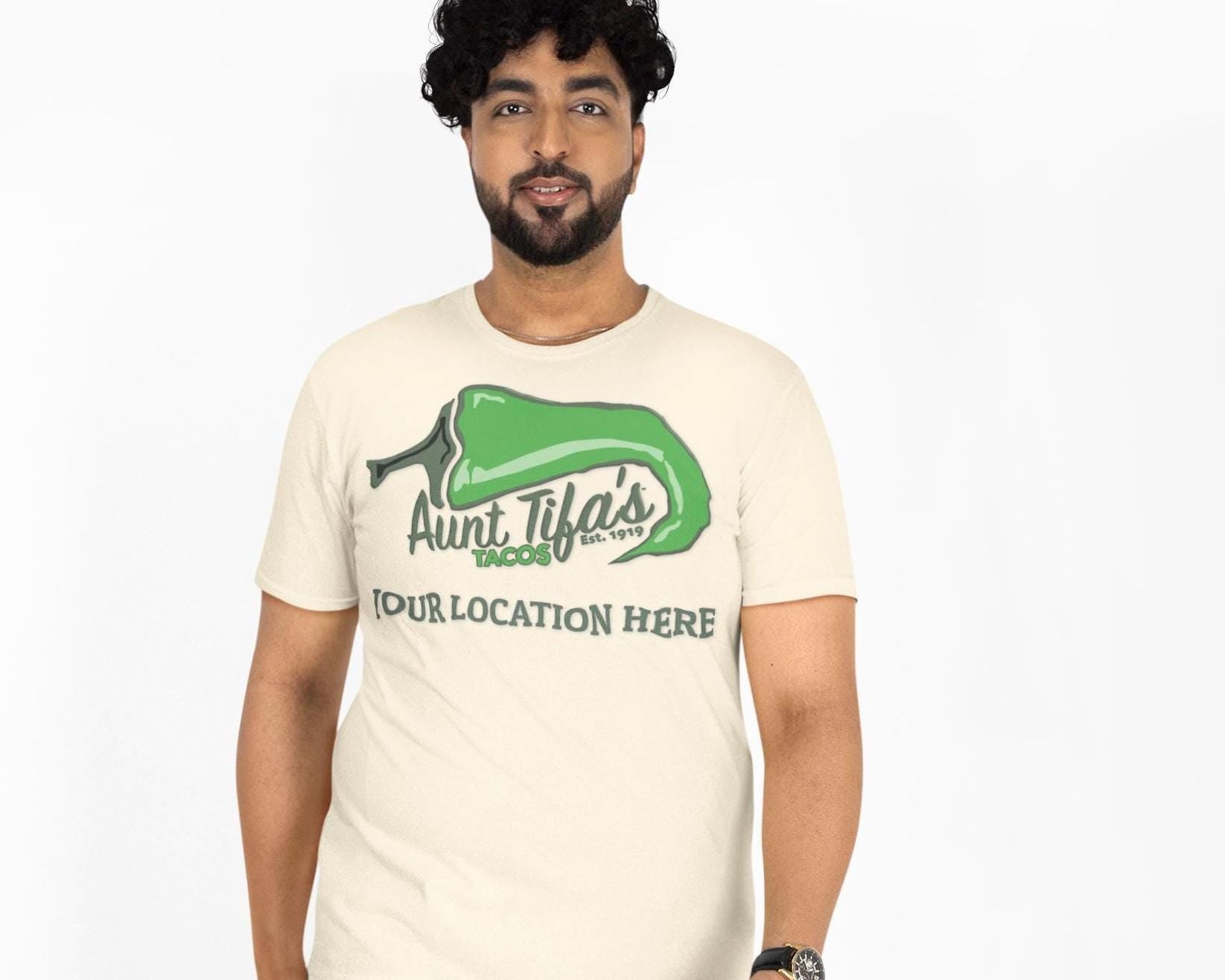 Aunt Tifa’s Tacos parody restaurant T-shirt with a customizable city name on white cotton fabric. Flat-front layout featuring the bold green-chili logo design. Satirical streetwear graphic for adults who love ironic humor and customizable parody merch.