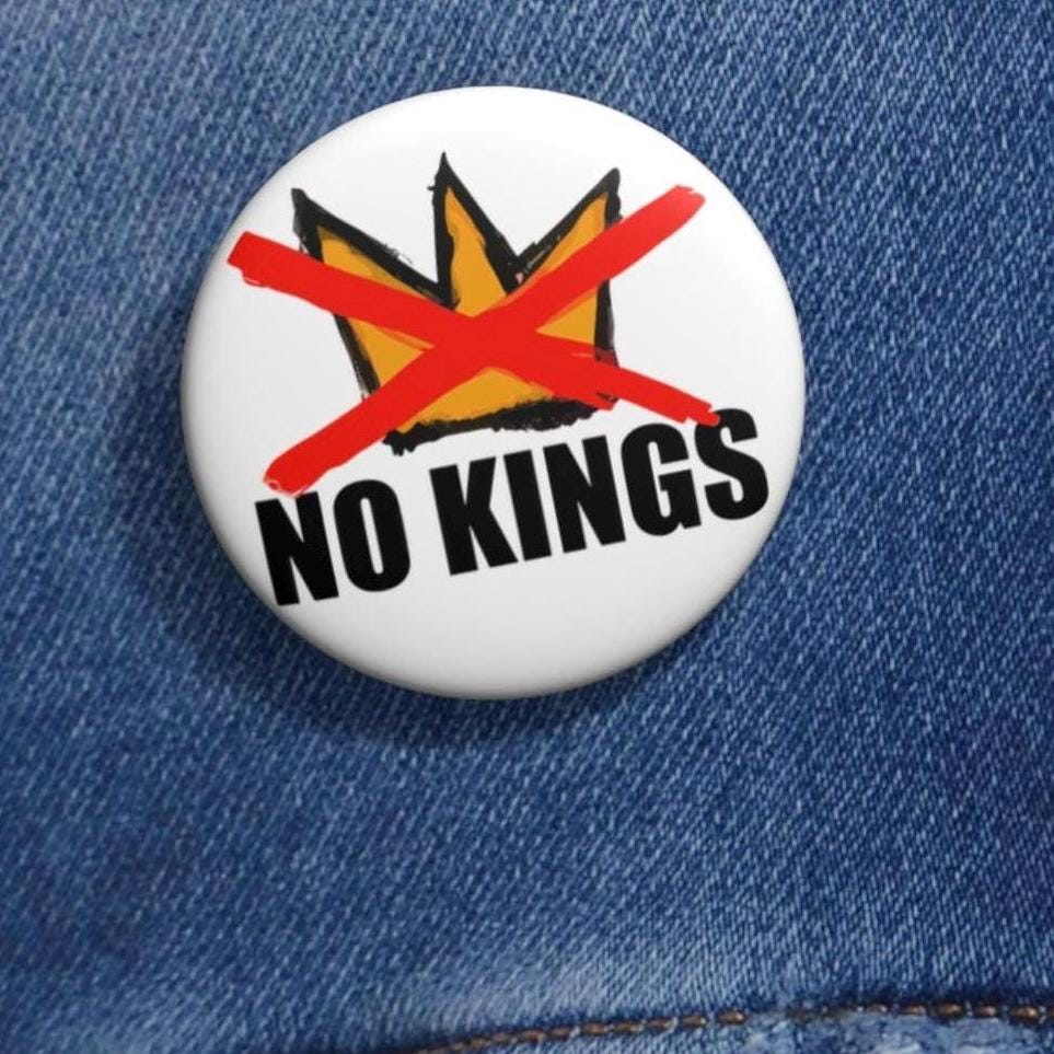 May include: A white button pin with the text "NO KINGS" in black, and a crossed-out gold crown. The pin is set against a blue denim jacket.