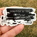 May include: A white sticker with black text that reads "EVERYTHING is FINE. TRUST your GOVERNMENT." The sticker has a distressed, typewriter-style font and design. The sticker is held in a hand.