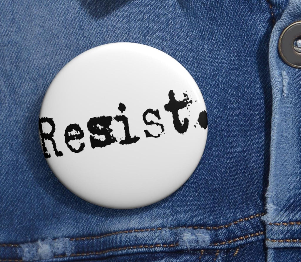 May include: A white button with the word "Resist" printed in black, typewriter-style font. The button is set against a blue denim background, with a button and stitching visible.