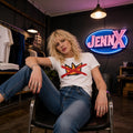 May include: A woman wearing a white t-shirt with a red and yellow graphic design, paired with blue jeans. The background features a neon sign that says "JennX". The setting appears to be a clothing store.